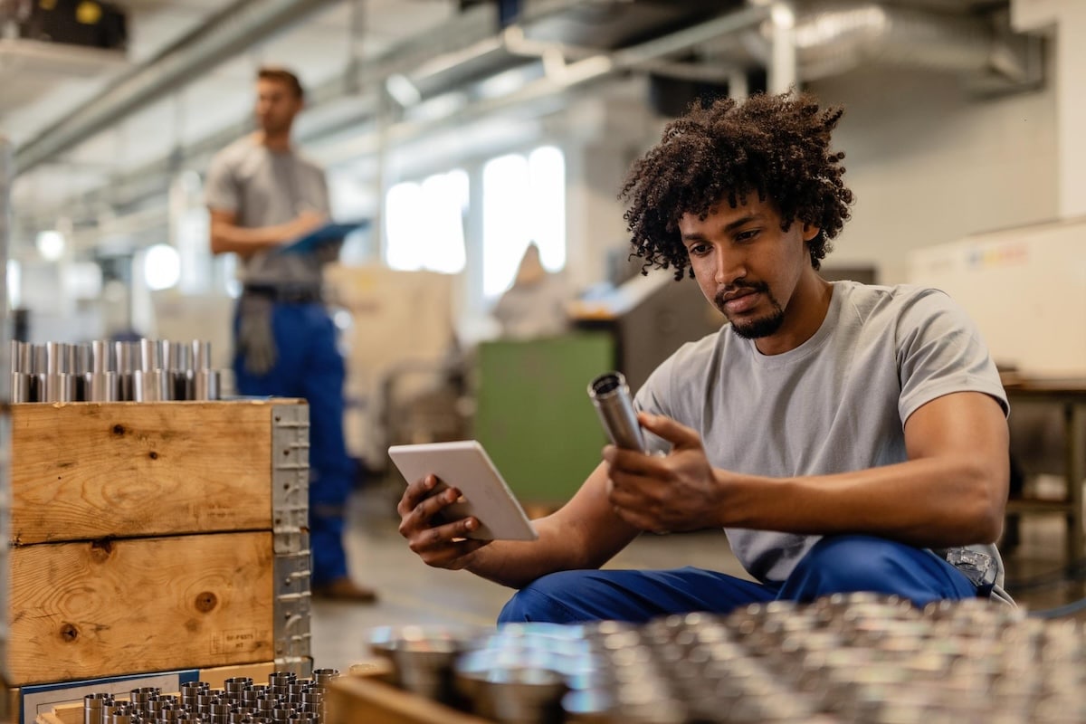 african-american-manual-worker-using-touchpad-while-examining-manufactured-stainless-steel-rods-factory